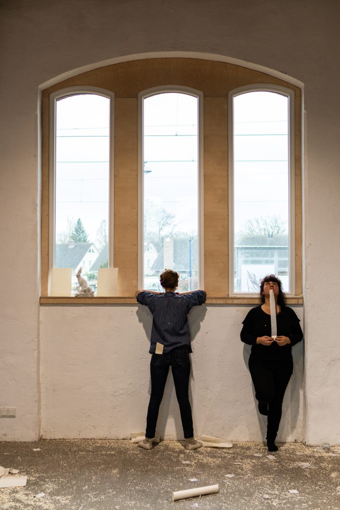 performance situtation: two women standing at a window looking in different directions, one out of the window, the other into the room