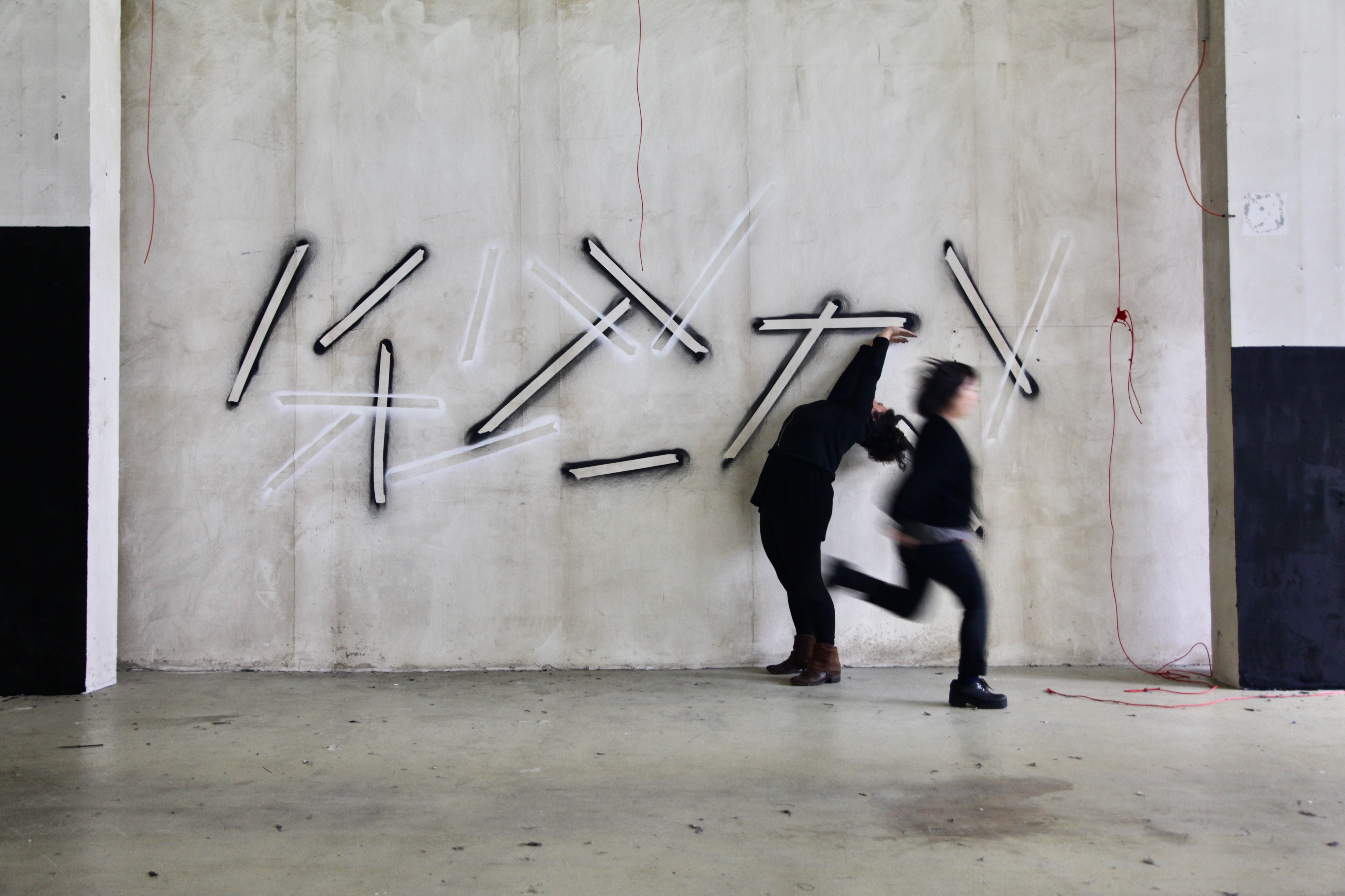 A concrete wall is sprayed with geometric elongated shapes. To the right and left of it stand concrete columns painted black in parts. In front of the wall are two people dressed in black. One is bending backwards. The other runs through the picture and is only blurred photographed.